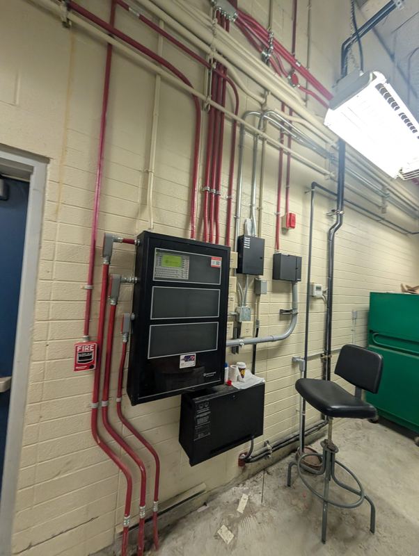 Fire alarm panel and power supply with surface-mounted red conduit runs on a masonry wall.